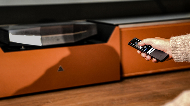 ine Person bedient einen Ultrakurzdistanz-Beamer mit einer Fernbedienung, der auf einem modernen Lowboard an der Wand platziert ist und so den Abstand zur störenden Lärmquelle im Heimkino maximiert.